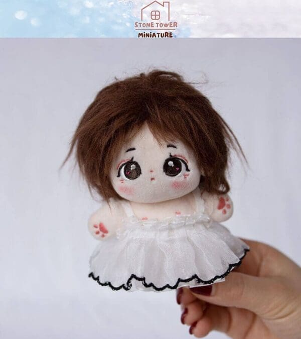 Cute plush doll with brown hair and large eyes, wearing a white dress, held in hand against a plain background.