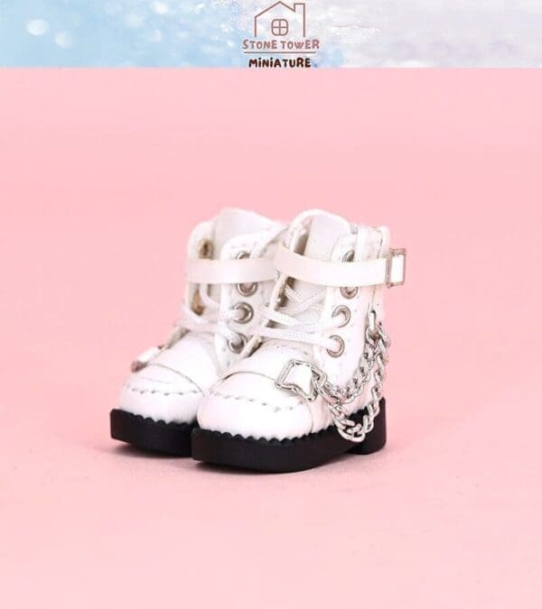 White miniature lace-up boots with silver chains on a pink background.