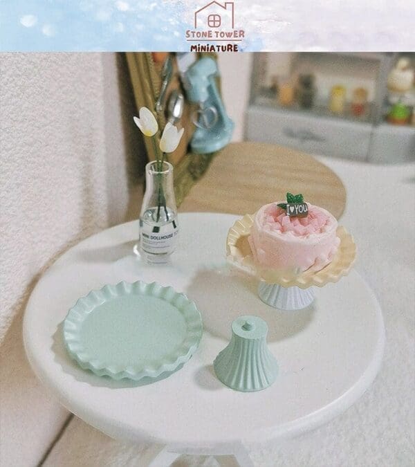 Miniature table with pink cake, green plates, and a vase with white flowers, creating a dollhouse scene.