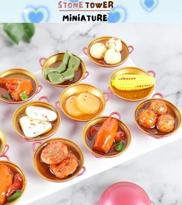 Miniature food in small golden pots, including corn, dumplings, and other colorful dishes on a white surface.